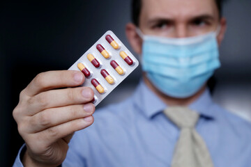 Worried man in protective face mask and office clothes looks at pills in his hand. Taking medication in capsules at work