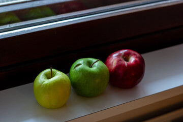 Red, green and yellow apples lie on the windowsill by the window.