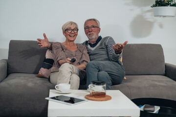 Surprised senior couple watching tv at home.
Excited senior couple is sitting embraced on couch in a living room and they are watching television together, movie or series online and relaxing