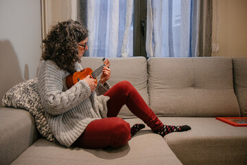 Mujer caucásica de mediana edad sentada en el sofá de su casa mientras està tocando el ukelele 