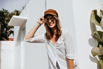 Fototapeta na wymiar Woman in high spirits coquettishly touches her wavy hair. Lady in cotton shirt and corduroy cap is smiling next to building in resort town