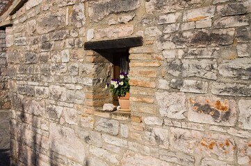 A small window under a wooden lintel decorated with a vase with white and violet flowers in a stone building of a medieval village (Marche, Italy, Europe)