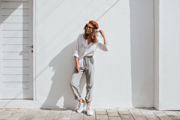 Stylish girl in gray pants and white cotton blouse posing near white wall. Photo of woman in cap...