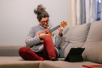 Mujer caucásica de mediana edad sonriendo sentada en el sofa con ropa casual aprendiendo  a tocar el ukelele por internet a traves de una tablet. - obrazy, fototapety, plakaty