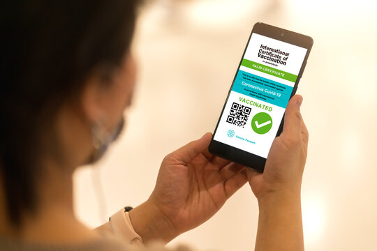 Smartphone displaying a valid digital vaccination certificate passport for COVID-19 in female's hand, public area background. Vaccination, disease immunity passport, health and safty travel concepts