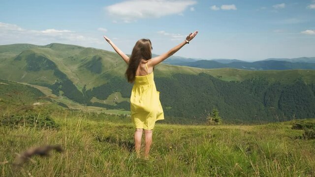 Back view of young happy woman traveler in yellow dress standing on grassy hillside on a windy day in summer mountains with outstretched arms enjoying view of nature.