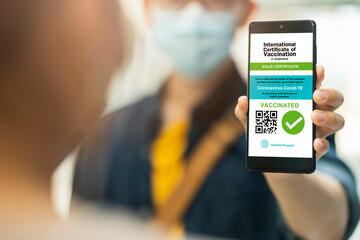 Smartphone displaying a valid digital vaccination certificate passport for COVID-19 in male's hand, public area background. Vaccination, disease immunity passport, health and safty travel concepts