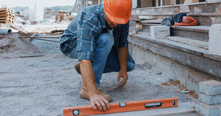 Builder in hard hat measuring ground with building level on construction site