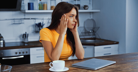 upset freelancer touching temples laptop near cup of coffee on table