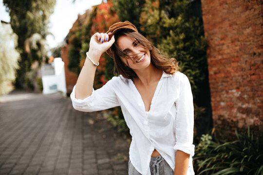 Wind Flinches Wavy Hair Of Young Woman Wearing Corduroy Hat. Shot Of Girl In White Blouse And Gray Shorts On Old Street With Bushes