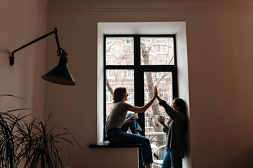 Side view of two girls drinking coffee near window. Indoor shot of friends holding cups of tea and showing high five sign.