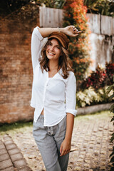 Obraz premium Picture of girl with short hair wearing brown cap, white blouse and linen trousers who raised her hand against background of brick building and bushes