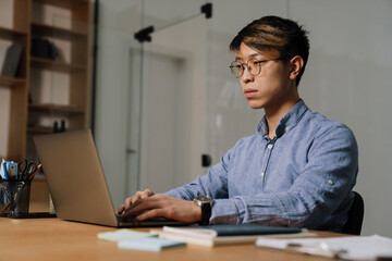 Fototapeta premium Focused asian guy working with laptop while sitting at table in office