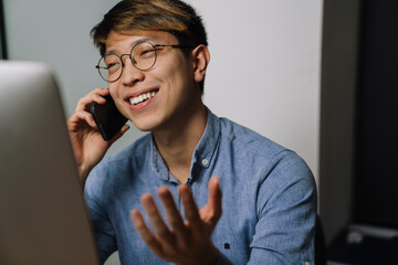 Joyful asian guy talking on cellphone while working with laptop