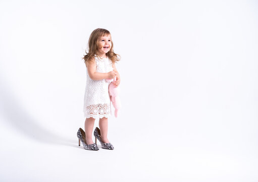 Funny little girl in white dress trying her mother shoes with heels on white background
