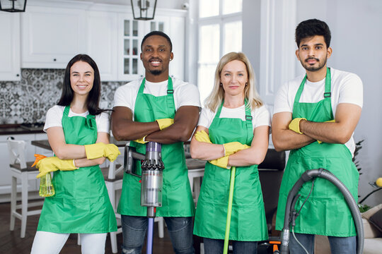 Confident mixed race people standing with crossed arms at apartment with modern interior. Team of cleaning service wearing green apron and yellow rubber gloves.
