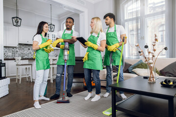 Multiracial team of cleaning service using digital tablet and talking during working process. Professional janitors in green apron using modern equipment at bright apartment.