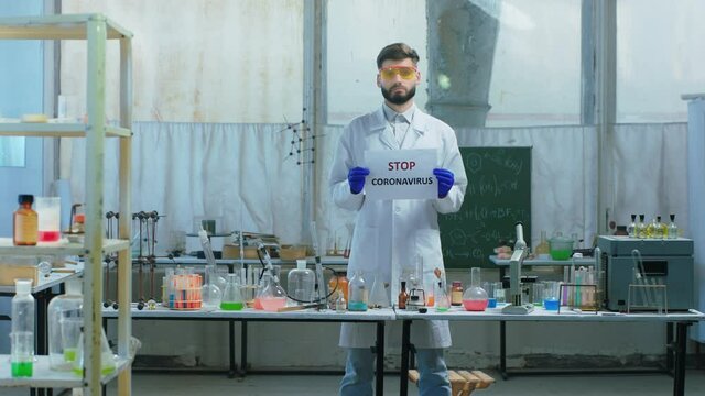 In a retro chemistry laboratory scientist man concentrated looking straight to the camera and holding a poster with Stop Coronavirus
