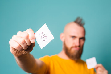 Bearded european man in yellow shirt isolated on turquoise background holding yes and no reminder afraid and shocked with surprise and amazed expression, fear and excited face.