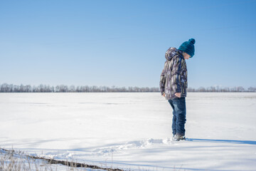 Fototapeta na wymiar A child in warm clothes walks in the fresh air in a snow field against a blue sky - measures the level of snow, stepping into the snow