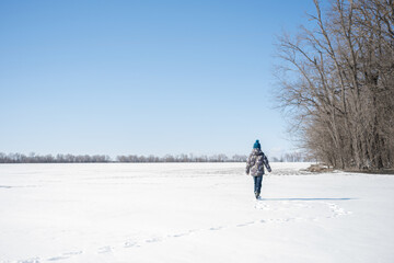 A child in warm clothes walks in the fresh air in a snowy field against the background of the blue sky and trees of the forest belt