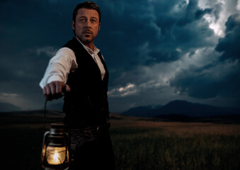Man in victorian clothing with lantern standing on prairie area.