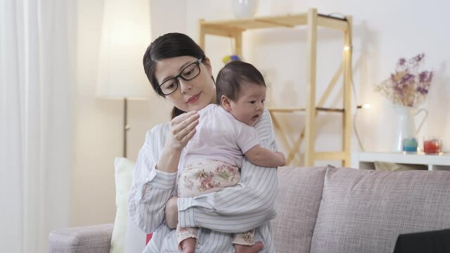 asian businesswoman is helping her newborn girl burp by holding her upright with gentle pats on the couch in the living room at home.