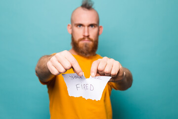 Bearded european man in yellow shirt isolated on turquoise background holding paper with you are fired tears up the paper with anger