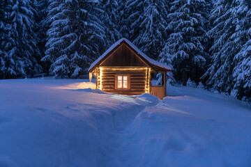 Fantastic winter landscape with glowing wooden cabin in snowy forest. Cozy house in Carpathian mountains. Christmas holiday concept