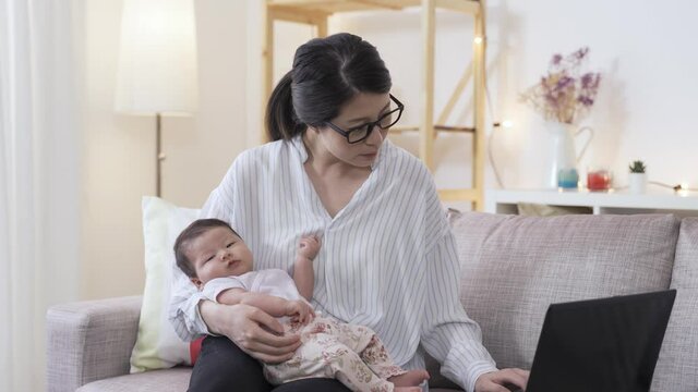 Korean working mother is telling the baby in her cuddle to be patient as she is replying email on the computer in the living room at home.