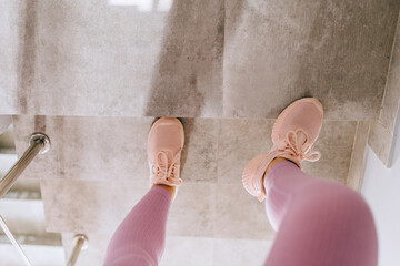 Top view of unrecognizable female legs in pink sportswear and sneakers go up in action on staircase in fitness studio or gym.