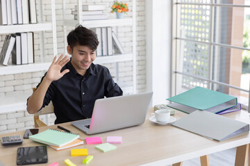 A young man greeting his friend online while using his computer to have an online conference during the work from home perios because of pendemic