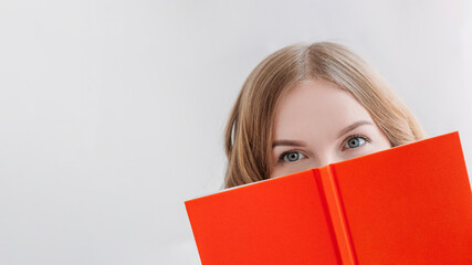 Portrait of a pretty young blond girl hiding behind an open orange book. Happy Woman studying or working at home