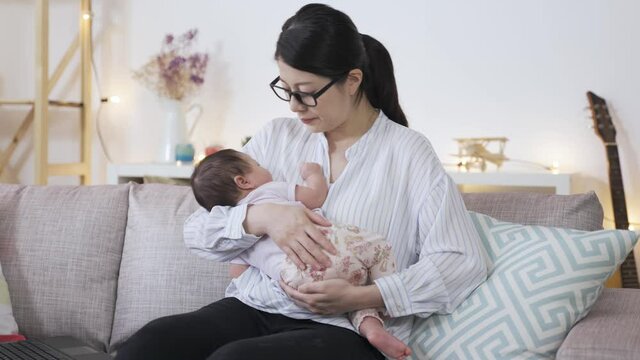 female employee is holding her baby daughter properly and gently patting on the bottom to coax her to sleep on the couch at home.