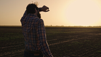 Male farmer making a rest of pushing wheelbarrow over farmland wearing straw hat plaid shirt rubber boots sunglasses at sunset