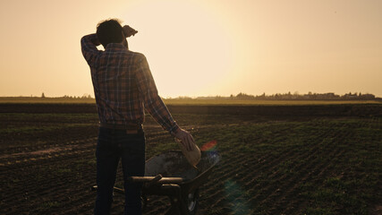 Male farmer making a rest of pushing wheelbarrow over farmland wearing straw hat plaid shirt rubber boots sunglasses at sunset
