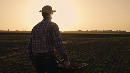 Male farmer pushing wheelbarrow over farmland wearing straw hat plaid shirt rubber boots sunglasses at sunset