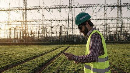Electrical engineer wearing a helmet and safety vest working with tablet near high voltage...