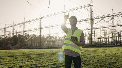 Electrical engineer wearing a helmet and safety vest working with tablet near high voltage electrical lines power station during sunset