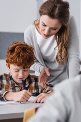 teacher pointing with hand while helping redhead schoolboy in classroom, blurred foreground
