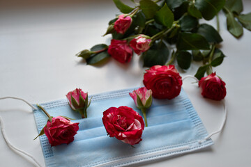 Fresh rosebuds on the medical face mask on the white table and bouquet of pink roses create delicate spring composition.