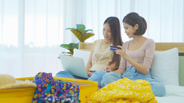 Happy Asian young women girls. people shopping online, using a credit card to pay money with a computer notebook laptop, suitcase luggage on bed in bedroom at home. Lifestyle.