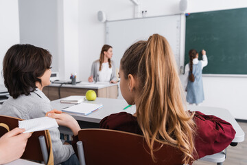 schoolgirl taking note from classmate near pupils and teacher on blurred background
