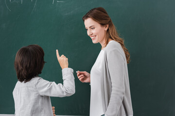 schoolboy pointing with hand near chalkboard and happy teacher