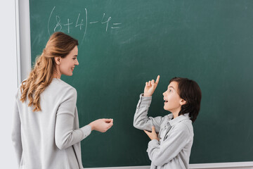 excited schoolboy pointing with finger at chalkboard near smiling teacher