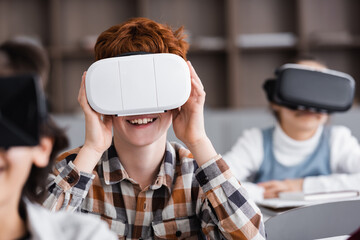 selective focus of cheerful redhead schoolboy gaming in vr headset near classmates