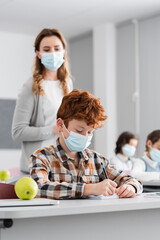 redhead boy in medical mask writing near apple and teacher on blurred background