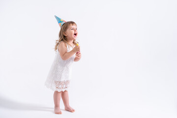 Funny child girl in white dress and birthday hat blowing in whistle on white background