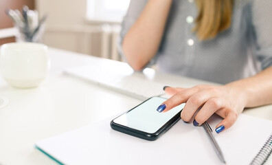 Fototapeta premium cropped shot of a young woman working from home using smart phone and computer, woman's hands using smart phone in interior, woman at home workplace using technology,