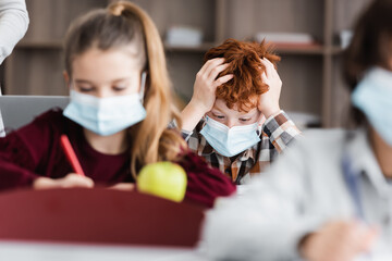 tired, redhead schoolkid in medical mask touching head near classmates on blurred foreground